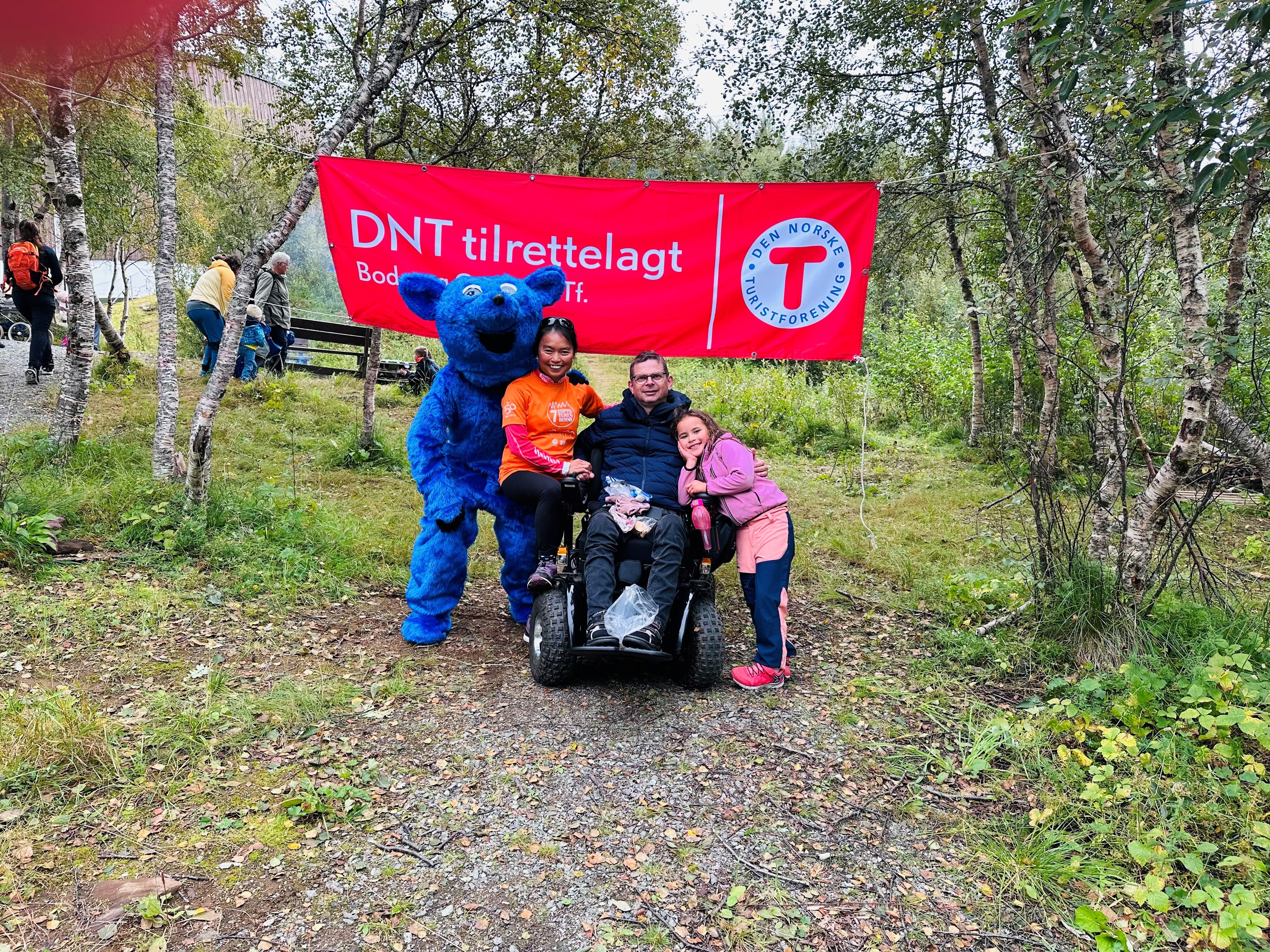En mann sitter i rullestol i skogen under et banner der det står "DNT tilrettelagt". Det står en blå rev og en kvinne på den ene siden og et barn på den andre siden av rullestolen.